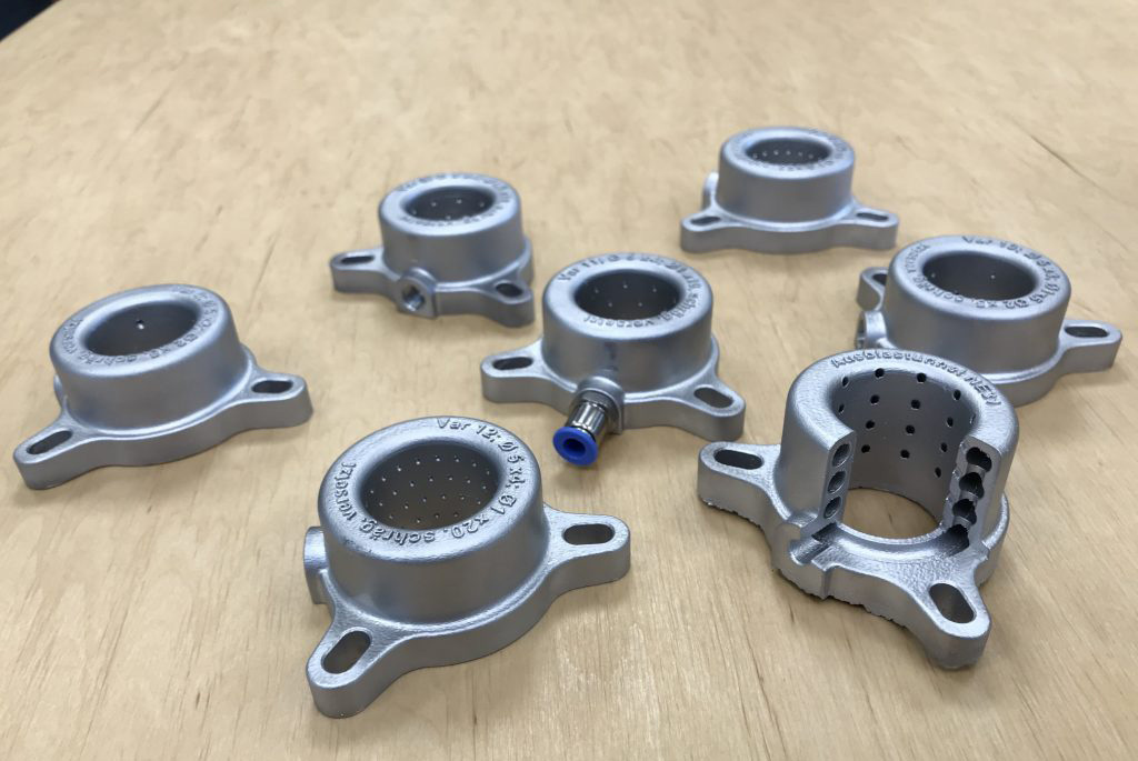 Ausblasdüse-min-2-1024×768 Sieben additiv gefertigte Ausblastunnel für CNC-Maschinen liegen auf einem Tisch. Ein Ausblastunnel ist zu einem Viertel aufgeschnitten, um die Luftbahnen besser sehen zu können